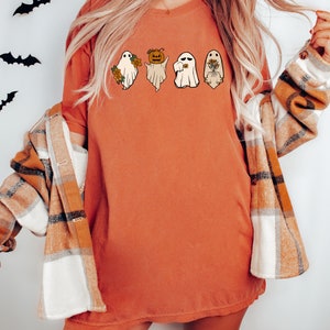 May include: A burnt orange t-shirt featuring a graphic of four ghosts. The ghosts are wearing different accessories, including a pumpkin, a flower, and a wreath.