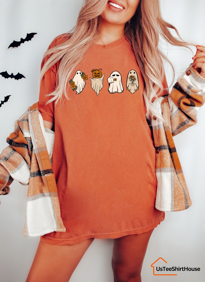 May include: A burnt orange t-shirt featuring a graphic of four ghosts. The ghosts are wearing different accessories, including a pumpkin, a flower, and a wreath.