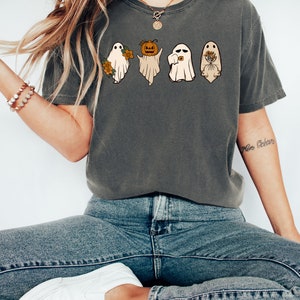 May include: A dark grey t-shirt featuring a graphic of three ghosts. The ghosts are wearing different accessories, including a pumpkin, a flower, and a bouquet of flowers. The text "More B&eacute;an" is visible on the right arm of the model wearing the shirt.