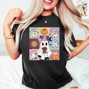 May include: Black t-shirt featuring a colorful, square graphic of cartoon ghosts with pink bows, holding books, and surrounded by pumpkins, flowers, and stars. The design has a retro, vintage aesthetic.
