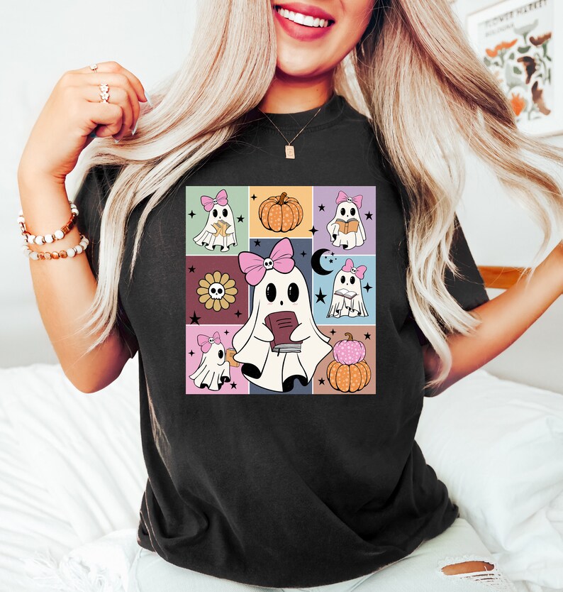 May include: Black t-shirt featuring a colorful, square graphic of cartoon ghosts with pink bows, holding books, and surrounded by pumpkins, flowers, and stars. The design has a retro, vintage aesthetic.