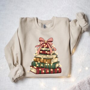 May include: Beige sweatshirt featuring a stack of books arranged in a Christmas tree shape. The books are in shades of red, green, and brown, adorned with a red and white plaid bow and string lights. A cozy, festive design.