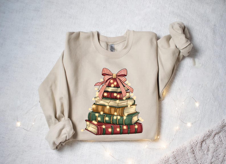 May include: Beige sweatshirt featuring a stack of books arranged in a Christmas tree shape. The books are in shades of red, green, and brown, adorned with a red and white plaid bow and string lights. A cozy, festive design.