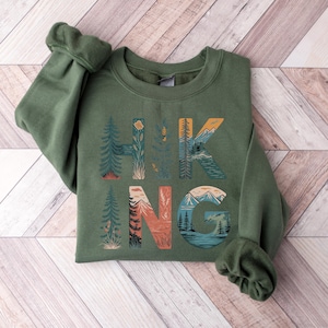 May include: A green sweatshirt with the word "HIKING" printed on the front. The letters are made up of illustrations of mountains, trees, and other nature scenes.