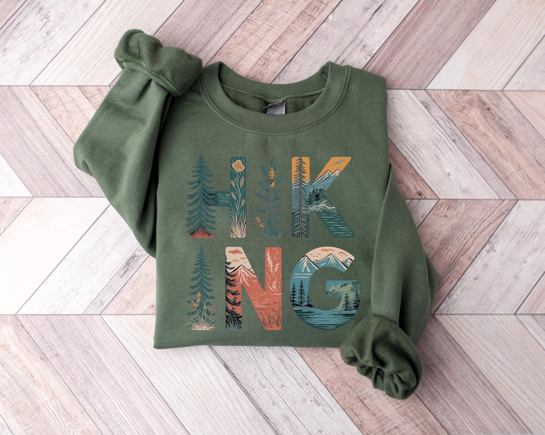 May include: A green sweatshirt with the word "HIKING" printed on the front. The letters are made up of illustrations of mountains, trees, and other nature scenes.