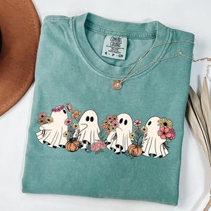 May include: A teal Comfort Colors t-shirt featuring a graphic of four cartoon ghosts with flowers and pumpkins. A gold necklace with a pendant is draped across the shirt. The shirt is folded.