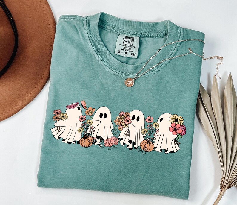 May include: A teal Comfort Colors t-shirt featuring a graphic of four cartoon ghosts with flowers and pumpkins. A gold necklace with a pendant is draped across the shirt. The shirt is folded.