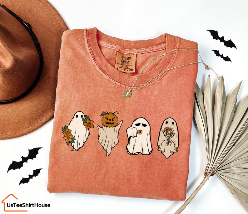 May include: A peach-coloured t-shirt with a graphic of three ghosts. The ghost on the left is holding flowers, the middle ghost is holding a pumpkin, and the ghost on the right is holding a bouquet of flowers. The text "COMFORT COLORS" is on the tag.