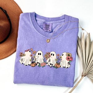 May include: A lavender Comfort Colors crew neck t-shirt featuring a graphic of four cartoon ghosts with flowers and pumpkins. The ghosts are white with black eyes. A gold necklace is draped across the shirt.