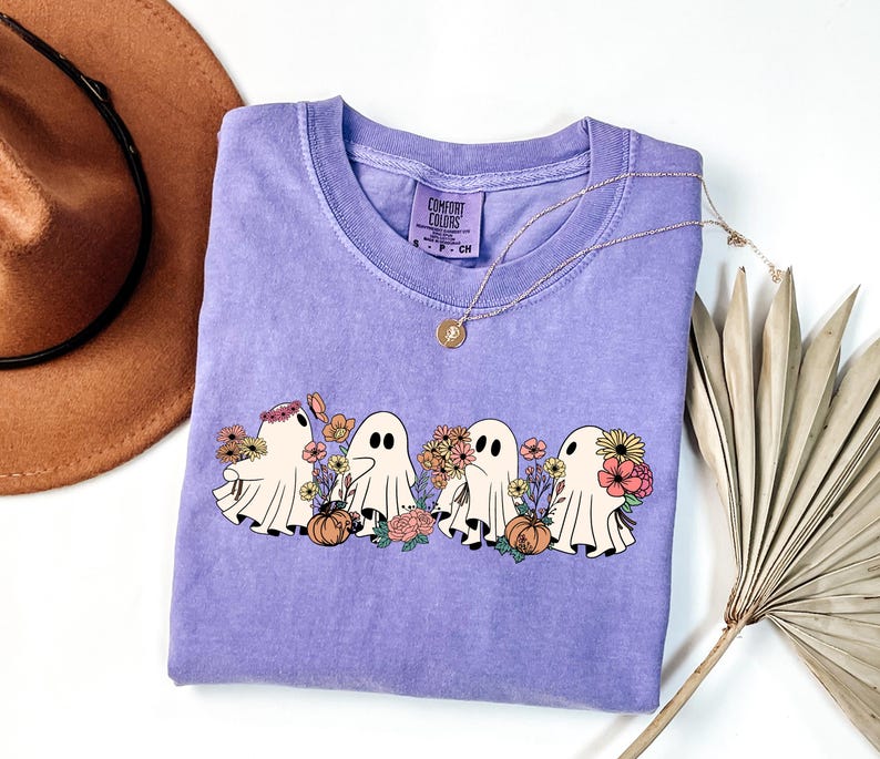 May include: A lavender Comfort Colors crew neck t-shirt featuring a graphic of four cartoon ghosts with flowers and pumpkins. The ghosts are white with black eyes. A gold necklace is draped across the shirt.