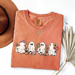 May include: A coral-colored Comfort Colors t-shirt featuring a graphic of four cartoon ghosts with floral arrangements and pumpkins. The ghosts have black eyes and are surrounded by colorful flowers. A gold necklace and a brown hat are also visible.