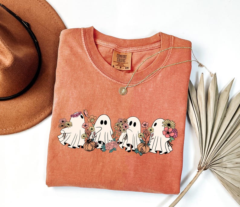 May include: A coral-colored Comfort Colors t-shirt featuring a graphic of four cartoon ghosts with floral arrangements and pumpkins. The ghosts have black eyes and are surrounded by colorful flowers. A gold necklace and a brown hat are also visible.