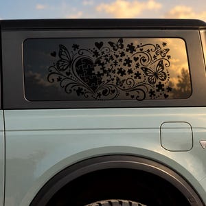May include: A black vinyl decal applied to the rear side window of a light-colored vehicle. The design features a heart shape composed of puzzle pieces, surrounded by intricate swirling flourishes, several butterflies, and small hearts.