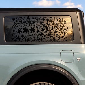 May include: A black vinyl decal applied to the rear side window of a light blue vehicle. The design features a central awareness ribbon composed of puzzle pieces, surrounded by intricate swirling flourishes, butterflies, and scattered puzzle pieces.