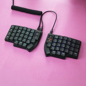 Peut inclure: Un clavier mécanique ergonomique divisé noir composé de deux moitiés distinctes reliées par un câble noir torsadé. Chaque partie présente une grille décalée de touches carrées avec lettrage blanc et un éclairage RVB intégré.