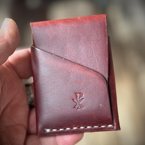May include: A dark red leather card holder featuring a diagonal pocket design and a stamped Chi-Rho symbol on the front. The bottom edge is secured with visible white stitching, and the leather surface displays a natural, slightly distressed texture.