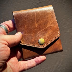 Op de afbeelding: Een vierkante bruinlederen portemonnee met een driehoekige klepsluiting die wordt vastgezet met een enkele messingkleurige metalen drukknoop. Het leeroppervlak vertoont een natuurlijke textuur met zichtbare krassen en kleurnuances.