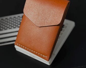 May include: A tan leather card holder featuring a pointed flap closure and visible light-colored stitching along the bottom edge. The wallet has a smooth texture and a structured, rectangular shape designed to hold cards securely in a compact form.