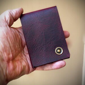 May include: A burgundy leather wallet with a textured finish. The wallet features a brass-coloured snap closure. The wallet is held in a hand against a blurred background.