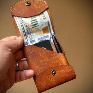 Puede incluir: Cartera de cuero marrón hecha a mano con cierre de botón a presión, que contiene efectivo, un bolígrafo negro y una tarjeta de crédito. La cartera tiene un diseño cosido y un pequeño logotipo.