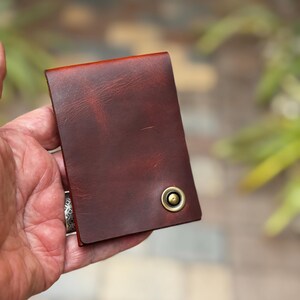 May include: A rectangular, dark reddish-brown leather wallet featuring a smooth, matte finish with visible natural grain textures. A single, circular, antique brass-colored metal snap fastener is positioned near the bottom right corner.