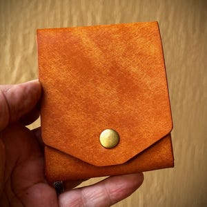 May include: A square-shaped wallet in a vibrant orange hue with a textured surface. The wallet features a pointed flap closure secured by a single, centered, circular brass-toned metal snap button, providing a simple and functional design.