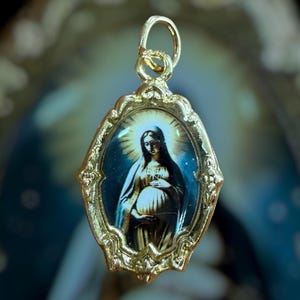 May include: An oval gold-toned pendant featuring a central image of a pregnant figure with a halo. The metal frame has an ornate, decorative border with intricate scrollwork details and a small circular bail at the top for attachment to a chain.