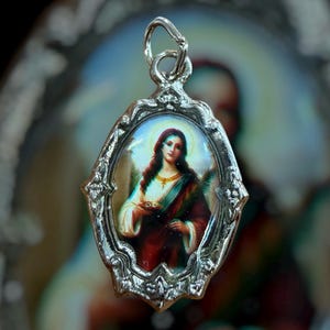 May include: An oval silver-toned metal pendant featuring a central color image of a woman with long dark hair, wearing a white garment and a red cloak, holding a small dish and a green palm frond, framed by an ornate, textured metal border.