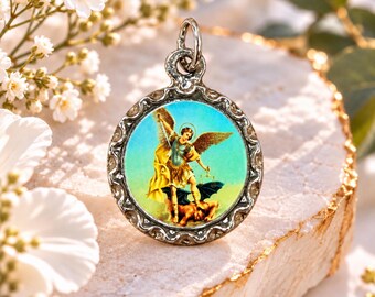 May include: A round silver-toned pendant featuring a colorful image of an angel with wings holding a sword and scales while standing over a figure. The pendant has a decorative scalloped edge and a small metal loop at the top for attachment.