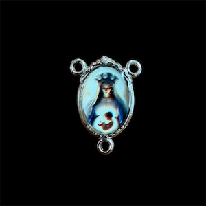 May include: A silver-toned metal rosary centerpiece featuring an oval-shaped inlay. The inlay displays a colorful depiction of a figure with a halo wearing a blue veil, holding a small child. The metal frame has three circular loops for attachment.