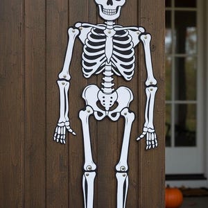 以下が含まれることがあります： ハロウィーン用の等身大の白と黒のスケルトン装飾。スケルトンは黒いアウトラインの白い切り抜きでできており、木のドアに取り付けられています。クラシックなハロウィーンの装飾です。