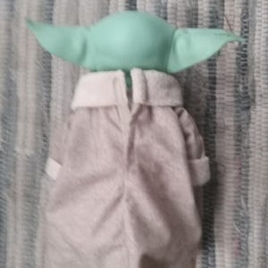 May include: A plush toy of a green baby alien wearing a gray hooded robe. The toy is facing away from the camera.