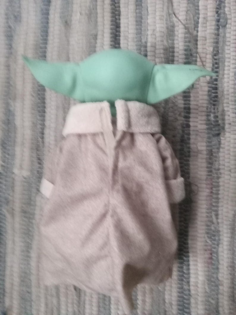 May include: A plush toy of a green baby alien wearing a gray hooded robe. The toy is facing away from the camera.