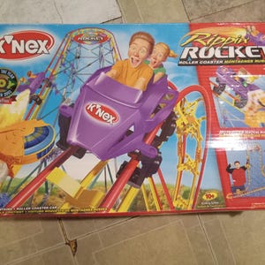 May include: K'NEX Rippin' Rocket roller coaster set box. The box features a purple roller coaster car with two excited figures. The set includes a roller coaster car and is for ages 10+. The box has the text "Toy of the Year 2003".
