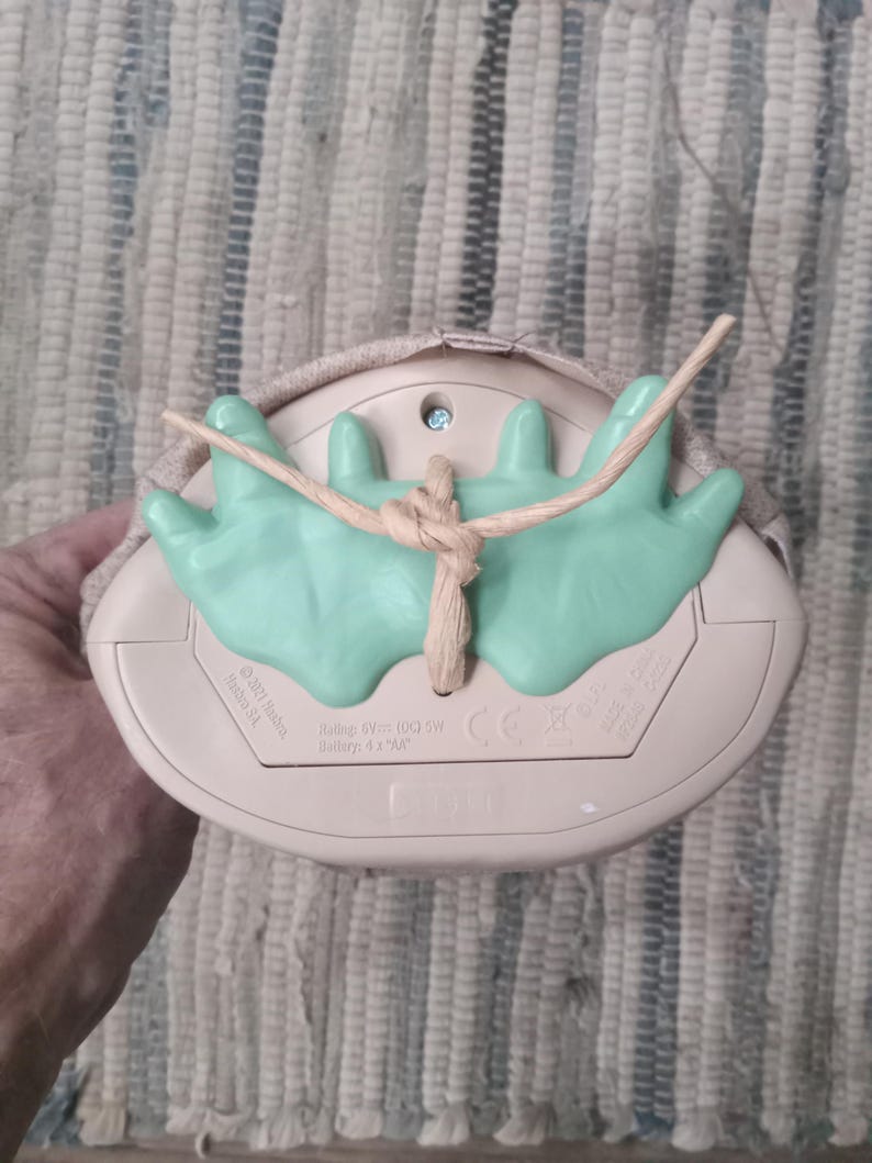 May include: A beige and green nightlight shaped like a mushroom with a string loop for hanging. The nightlight has a small light bulb and a battery compartment on the bottom. The text on the bottom reads "Rating 6V (DC) 5W Battery: 4.5V AA" and "CE".