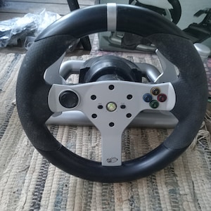 May include: A black and gray Xbox 360 racing wheel with a silver center. The wheel has a textured grip and colored buttons. The brand name Mad Catz is visible on the wheel.