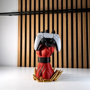 May include: A red sculpted hand stand featuring a black wrist cuff and gold-colored bullet accents at the base, holding a white and black wireless video game controller. The controller features standard directional buttons, analog sticks, and symbols.