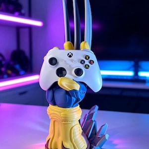 May include: A controller stand shaped like a blue and yellow gloved hand with three long silver claws extending upward. The hand holds a white wireless game controller featuring a black directional pad, two analog sticks, and four colored buttons.