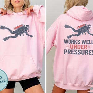 A woman wearing a pink hoodie with a graphic of a scuba diver and the text "WORKS WELL UNDER PRESSURE!" printed on the back.