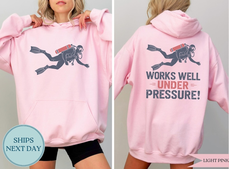 A woman wearing a pink hoodie with a graphic of a scuba diver and the text "WORKS WELL UNDER PRESSURE!" printed on the back.