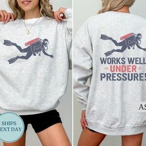 A woman wearing a gray sweatshirt with a graphic of a scuba diver and the text "WORKS WELL UNDER PRESSURE!" printed on the back.