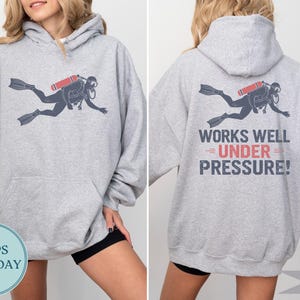 A woman wearing a grey hoodie with a graphic of a scuba diver and the text "WORKS WELL UNDER PRESSURE!" printed on the back.