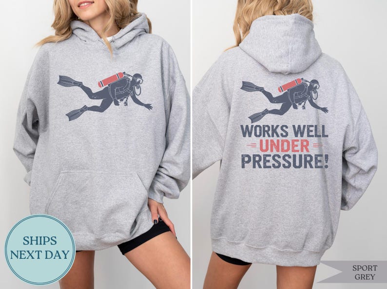 A woman wearing a grey hoodie with a graphic of a scuba diver and the text "WORKS WELL UNDER PRESSURE!" printed on the back.