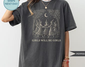 May include: Dark gray t-shirt featuring a graphic of five women in witch hats dancing in a circle under a crescent moon and trees. The text "GIRLS WILL BE GIRLS" is printed below the graphic. The shirt has a relaxed fit.