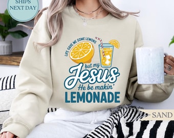 May include: Sand-colored sweatshirt with the text "Life gave me some lemons but my Jesus He be makin' LEMONADE" and a graphic of a lemon and a glass of lemonade. The sweatshirt has a crew neck and long sleeves.