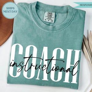 May include: Seafoam green t-shirt with the word "COACH" in large white block letters and the word "instructional" in a cursive font underneath. The shirt is made of a soft material and has a crew neck.
