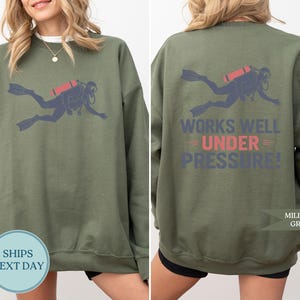 a woman wearing a green sweatshirt with a graphic of a scuba diver and the text "Works well under pressure!" printed on the back.