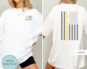 May include: White t-shirt with a small American flag design on the front and a larger flag design on the back. The back design includes the text "LAST NAME" in yellow. The flag features gray stripes and a yellow stripe.