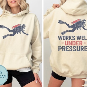 May include: Sand-colored hoodie featuring a diver graphic on the front and back. The diver is depicted in navy blue with a red oxygen tank. The back of the hoodie has the text "WORKS WELL UNDER PRESSURE!"