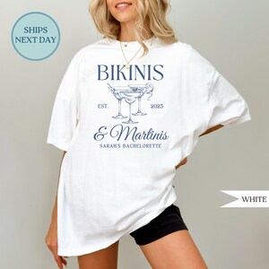 May include: White t-shirt with a navy blue design featuring the words "Bikinis & Martinis" and martini glass illustrations. The text "Sarah's Bachelorette" is also included. The shirt is a casual style.